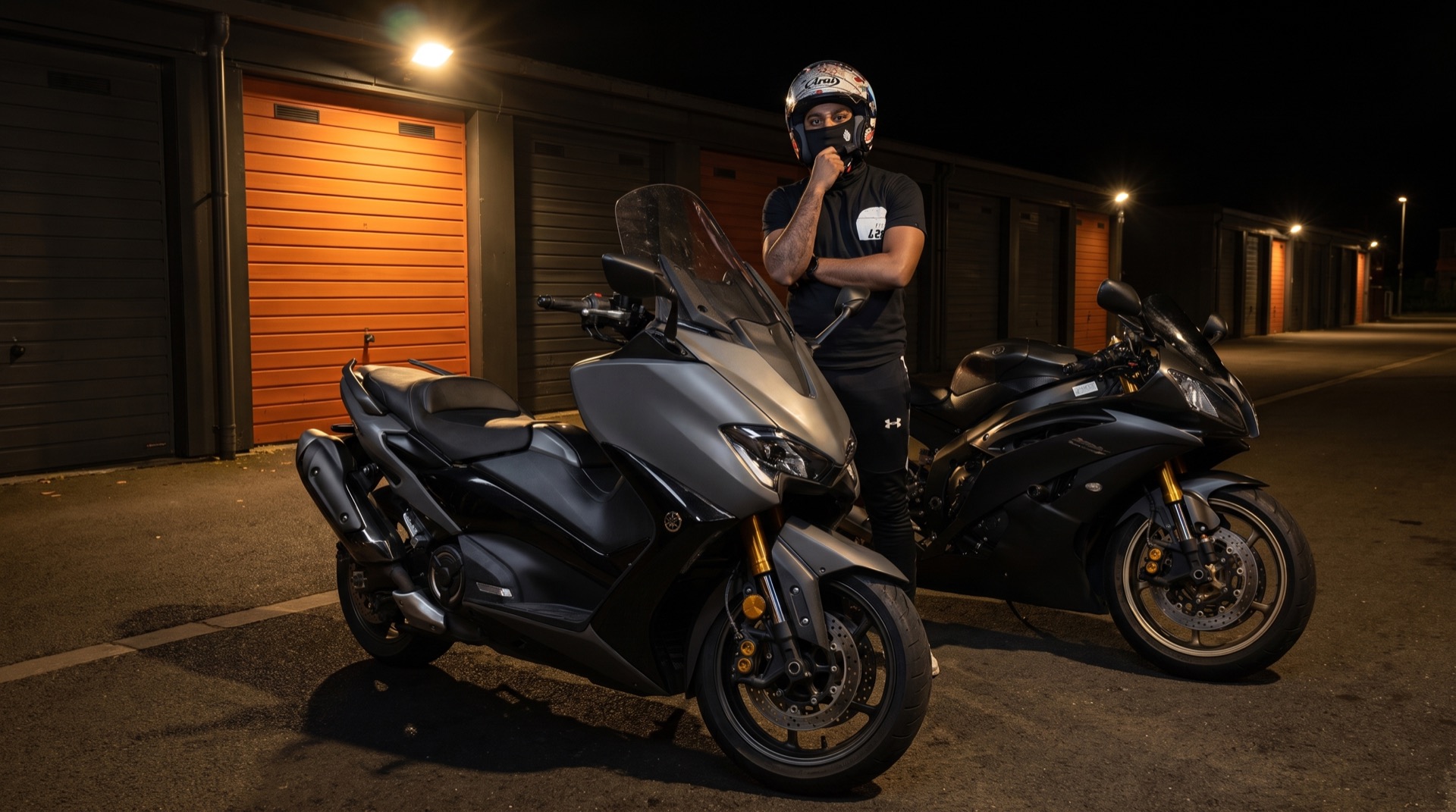 Un maxi-scooter et une sportive garés de nuit devant les box, rideau orange éclairé, le pilote en tenue noire entre les deux machines.